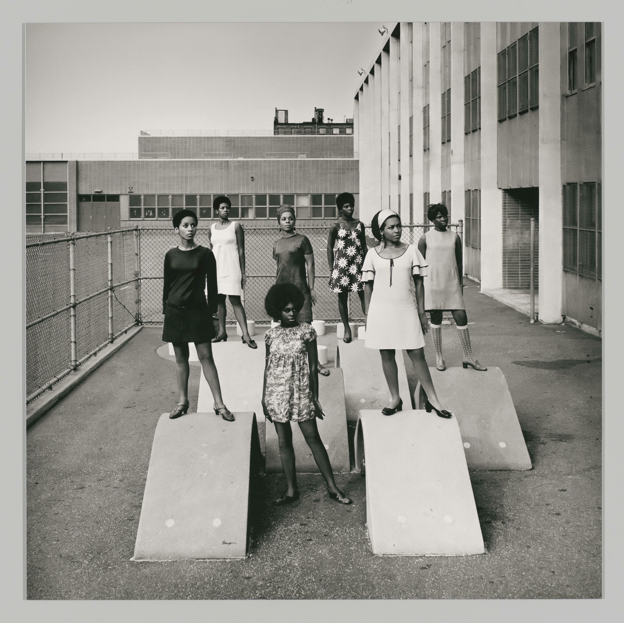 A black and white photograph depicting seven girls of varying ages standing on concrete pedestals in a rooftop setting, with a chain-link fence and industrial buildings in the background. The girls are dressed in a variety of outfits, including dresses and skirts, and most are looking directly at the camera with neutral expressions.