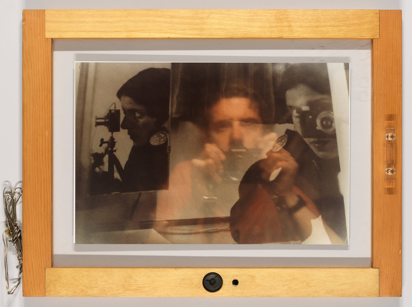 A framed photograph on a wall showing a layered image of three people, with the central figure holding a transparent cup to their mouth. The person on the left is in profile, looking at a device, and the person on the right is holding a camera to their eye. The frame is wooden with a visible lock at the bottom.