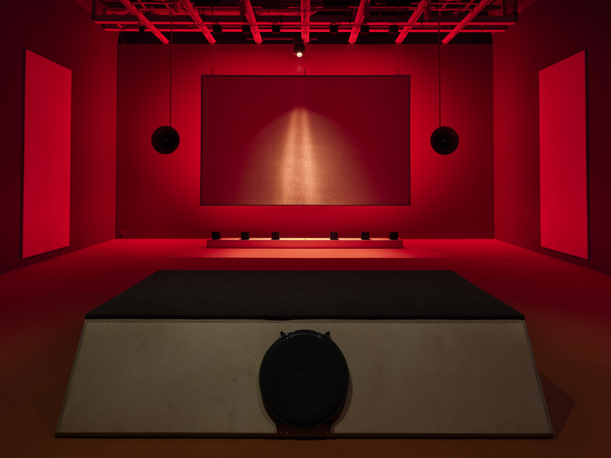 A room lit with red lights, featuring a raised platform, speakers, and large blank panels on the walls.