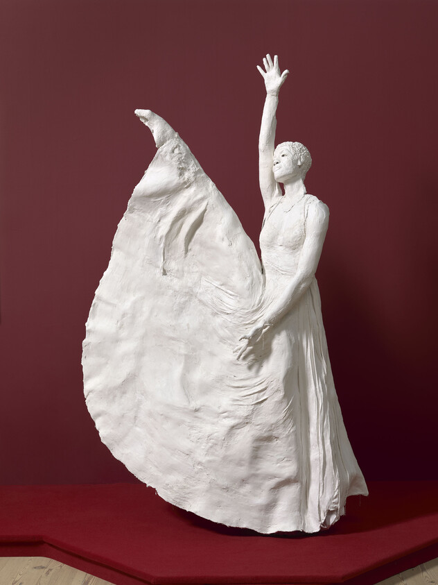 White sculpture of a woman in a flowing dress, one arm raised, against a dark red background.