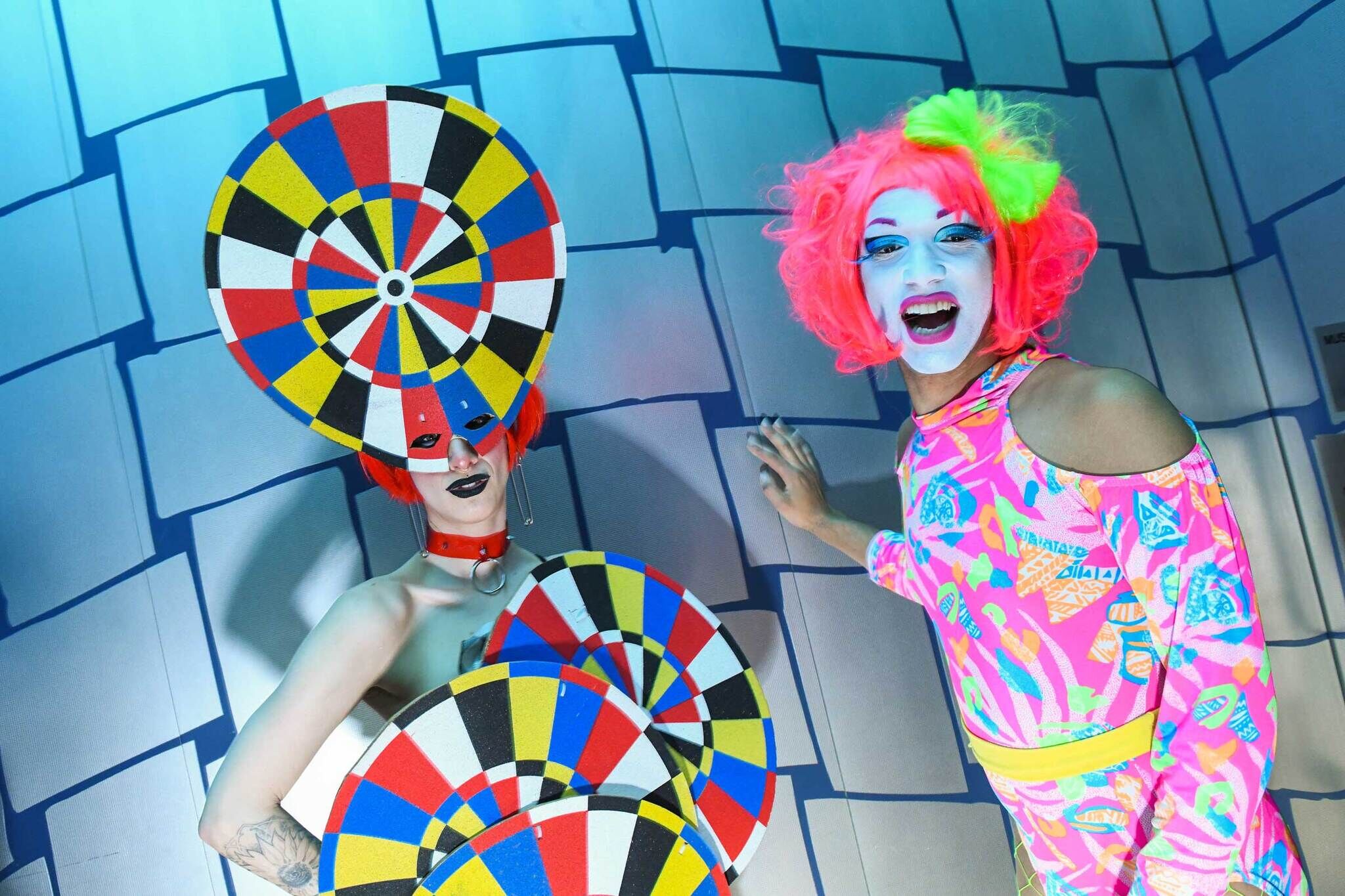 Two people in colorful, bold costumes and wigs pose against a blue tiled wall, smiling and playful.
