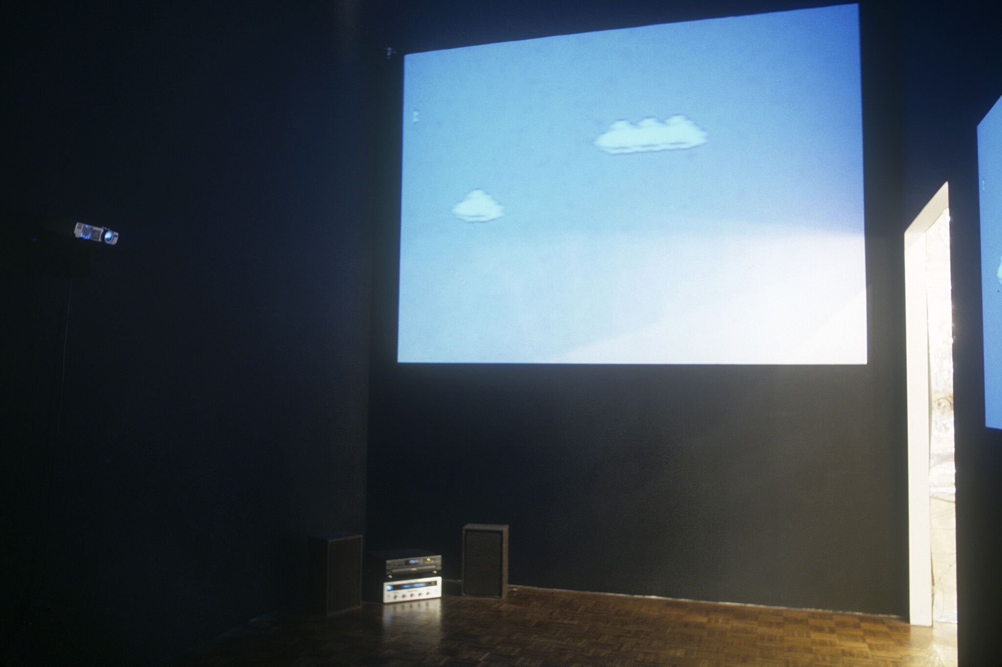 A gallery with dark walls displaying a projection of a blue sky with pixelated clouds.