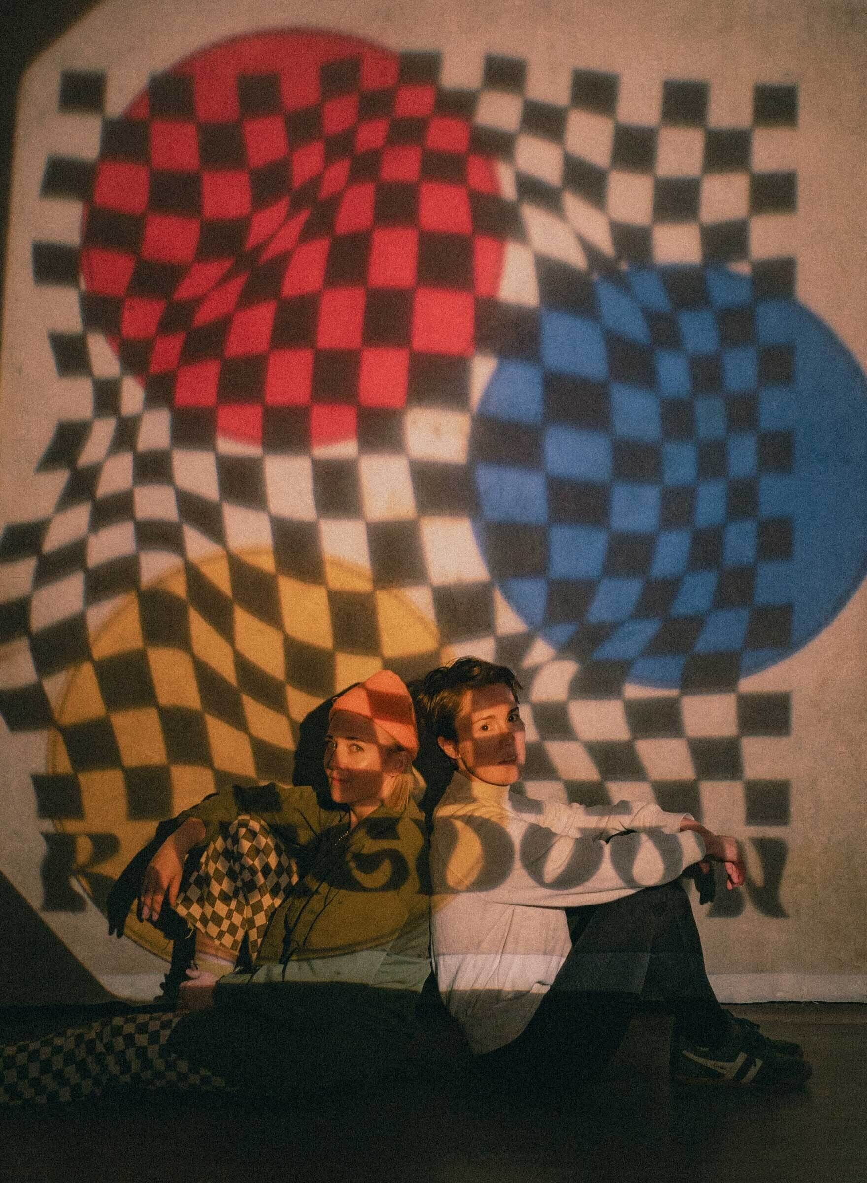 Caroline Shaw and Danni Lee Parpan sitting back to back. A checkered pattern with red, blue, and yellow circles is projected on them and against the wall behind, with the words "Ringdown" overlaid.