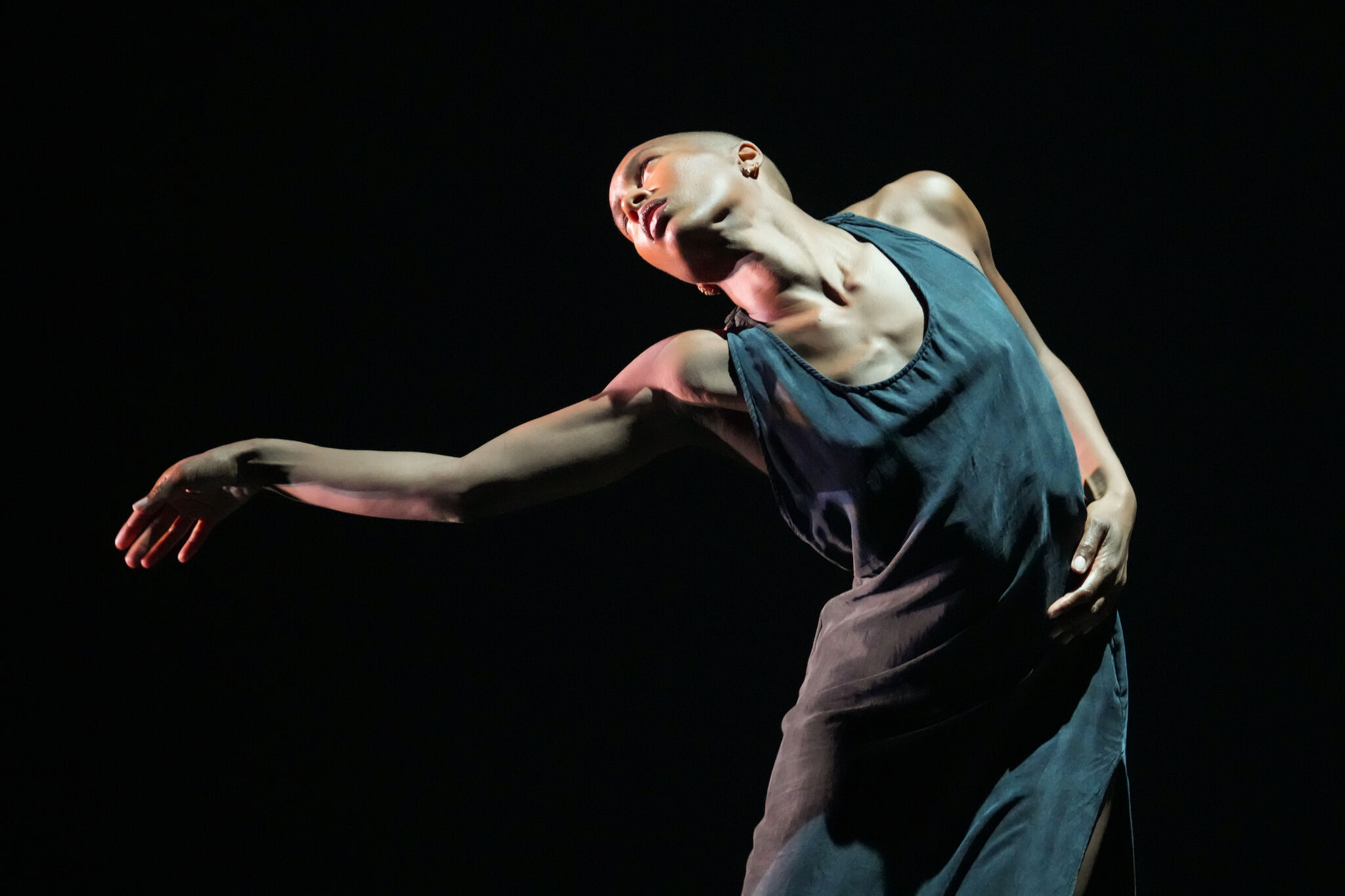 A dancer in a flowing dress strikes a graceful pose against a dark background, with one arm extended and head tilted back.