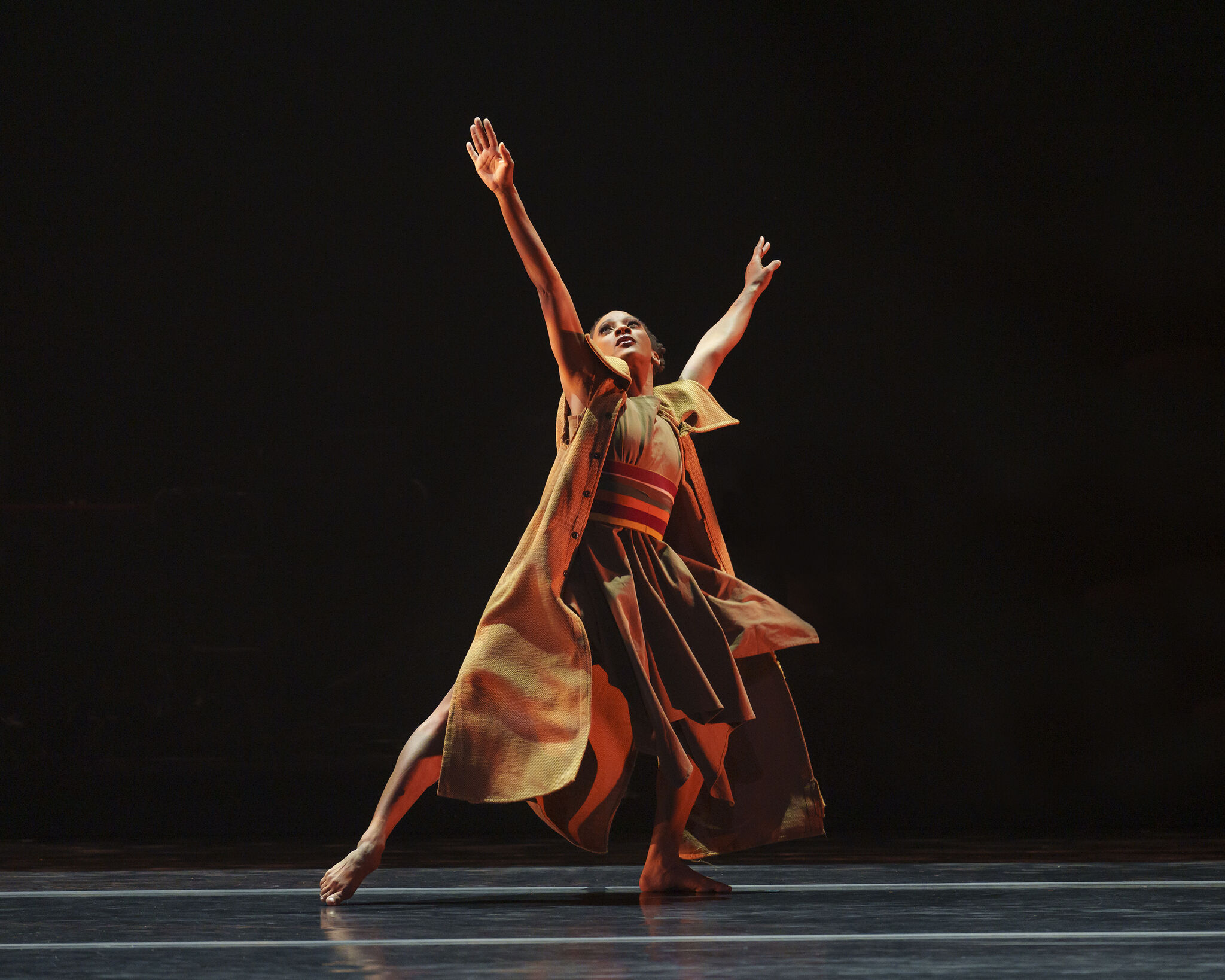 A dancer in a flowing dress strikes a dramatic pose on stage, arms reaching upward against a dark background.
