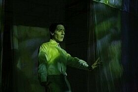 A man in a white shirt with their arm outstretched, half shrouded in shadow. Everything has a green tint.