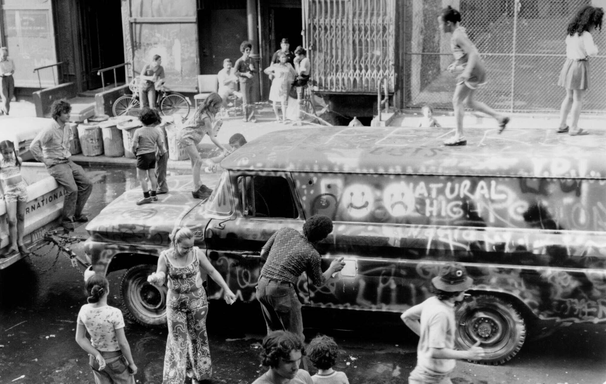 Black and white photo of people painting and playing on a graffiti van on the streets.