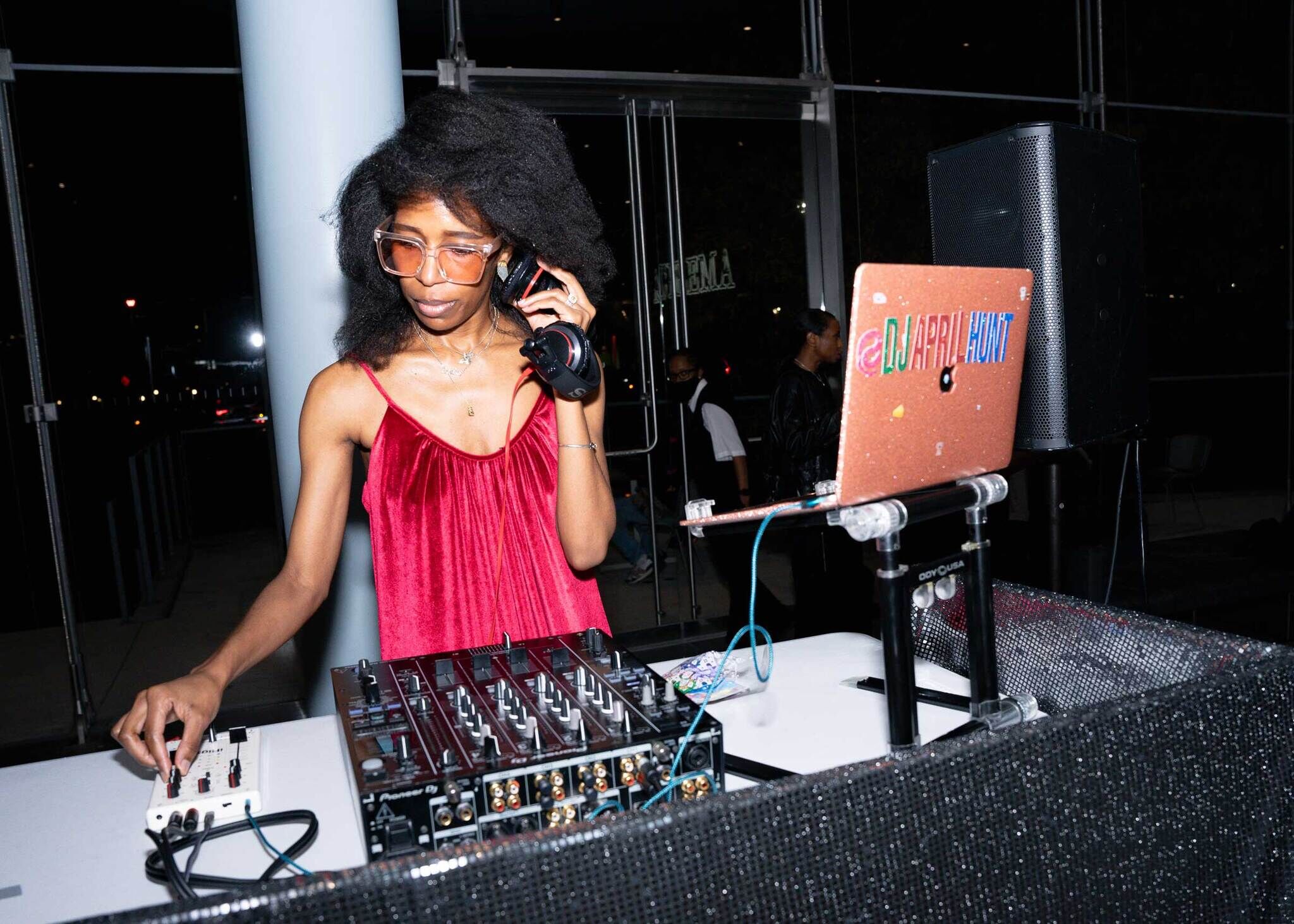 A DJ wearing pink dress and glasses adjusting equipment in the booth.