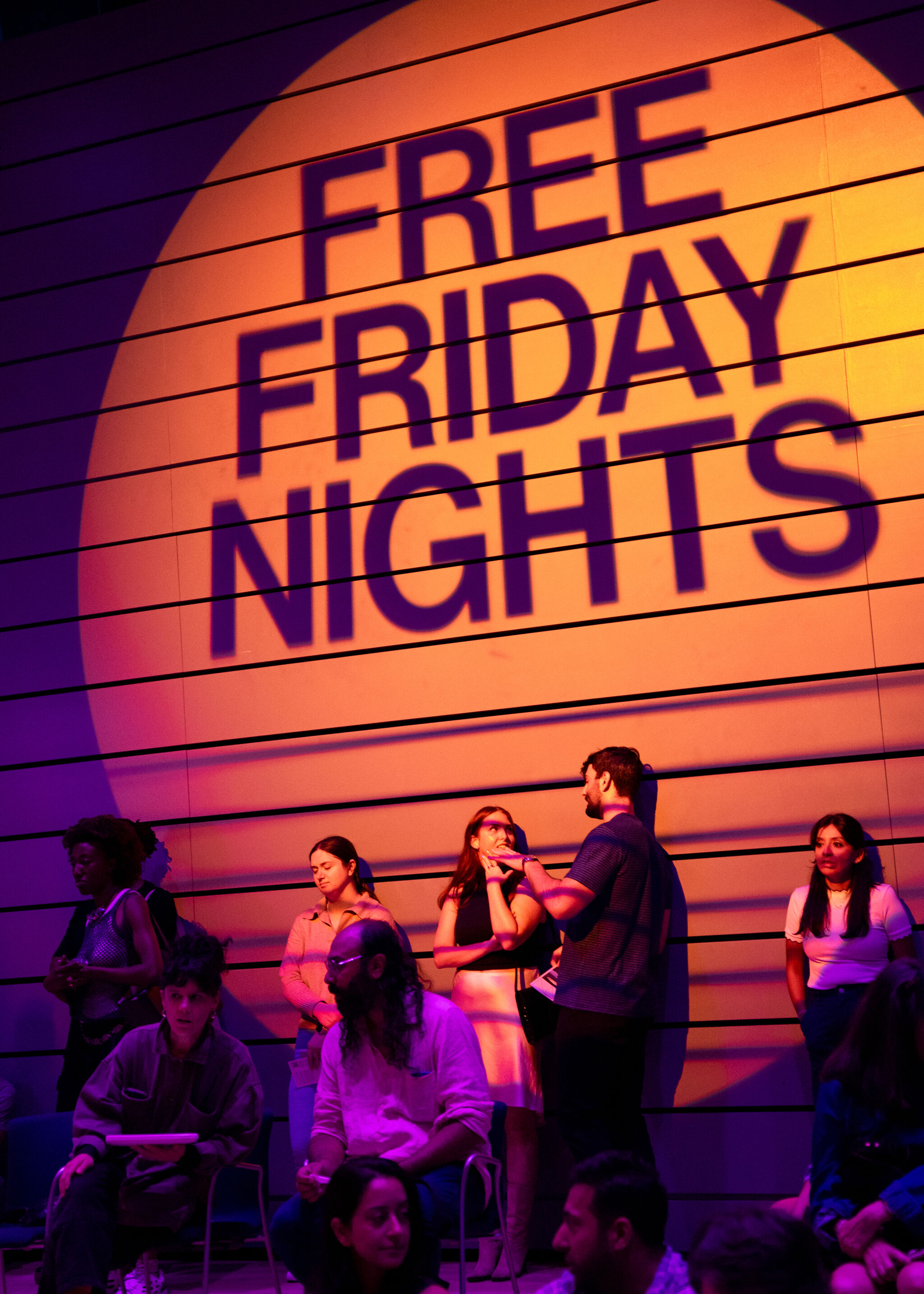 The words "Free Friday Nights," seen within a sunset-colored orb, are projected onto a wall, with visitors leaning on it and conversing.