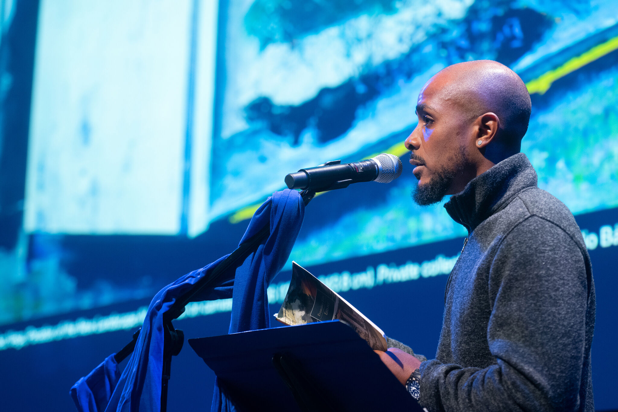 A person stands at a podium and microphone with a bright blue illuminated screen behind. 