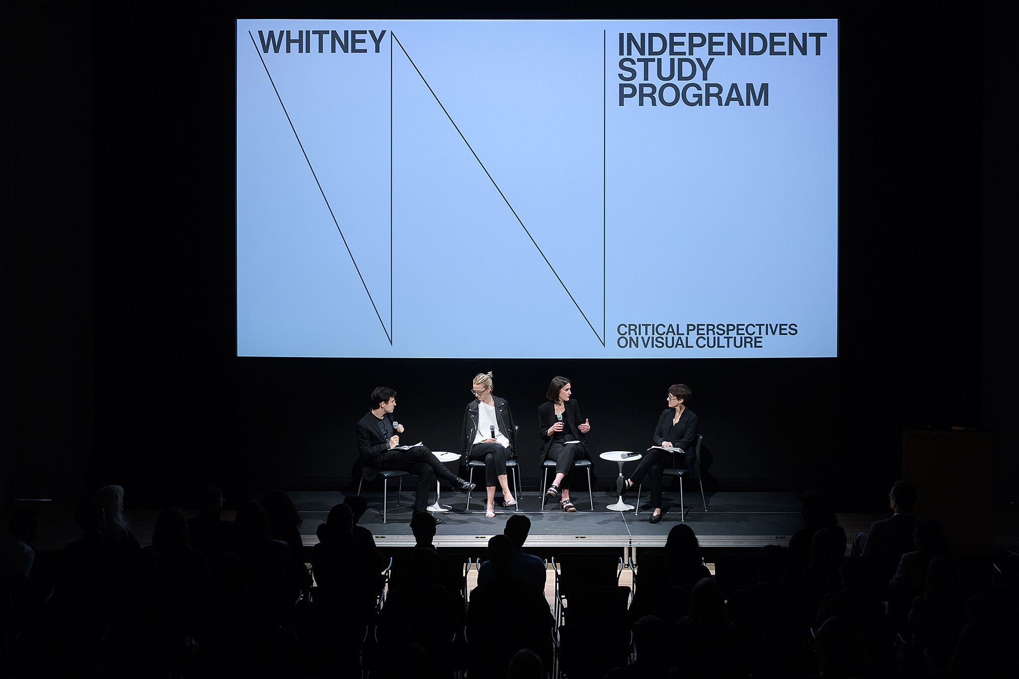 Four people sit on a stage, with a screen lit up behind them with the Whitney Museum logo