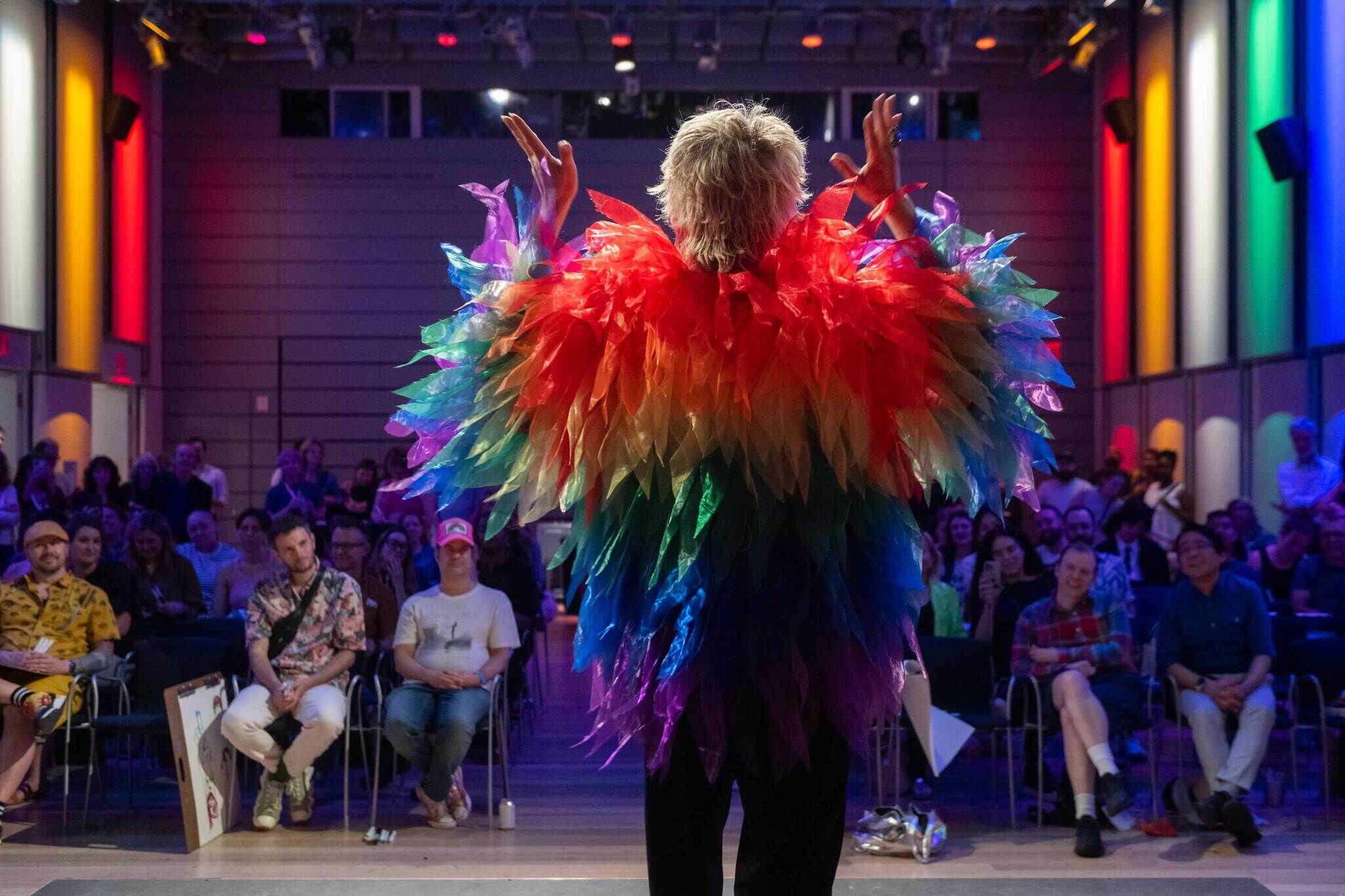 Person wearing a bright rainbow feathered costume stands on stage, facing an audience seated in a colorful room.