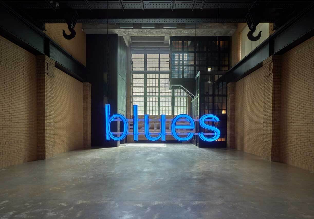 Blue neon sign that reads, "blue," in front of a window in a brick-walled, loft-like space.