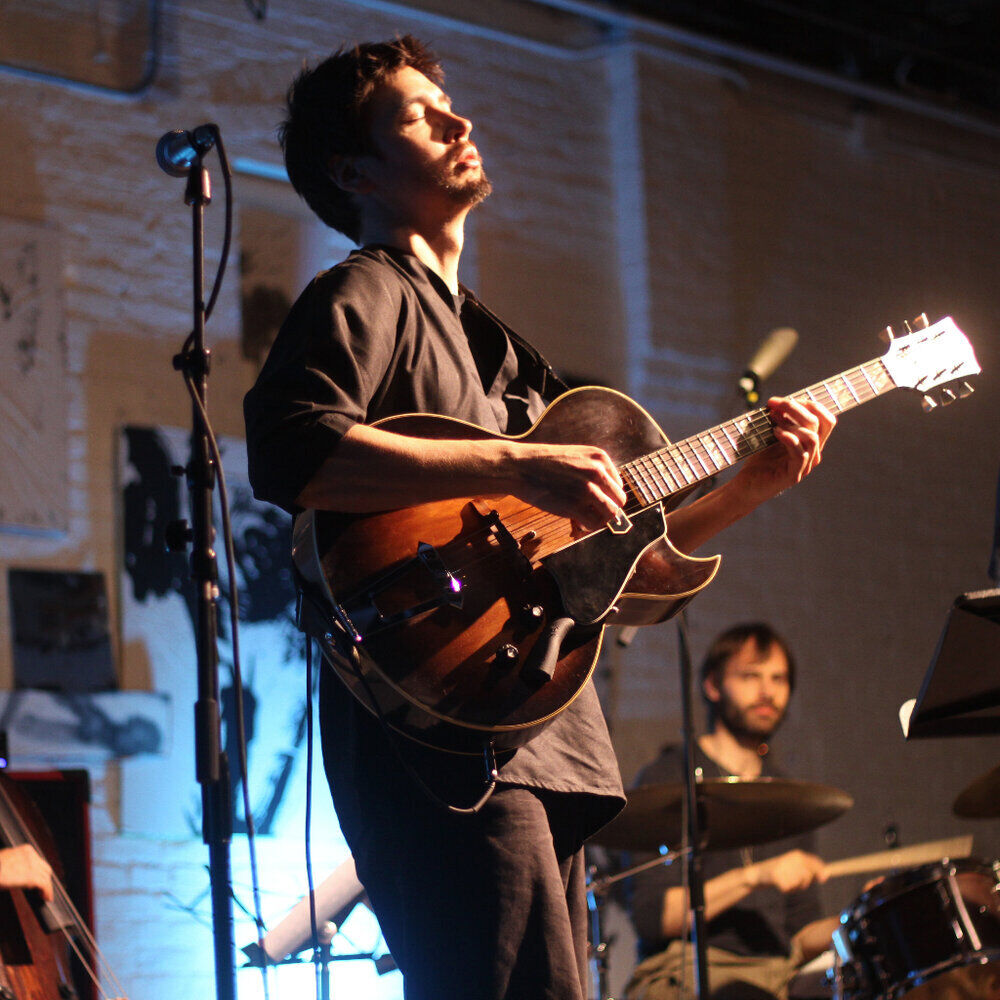 Man playing an electric guitar on stage with eyes closed, drummer performing in the background.