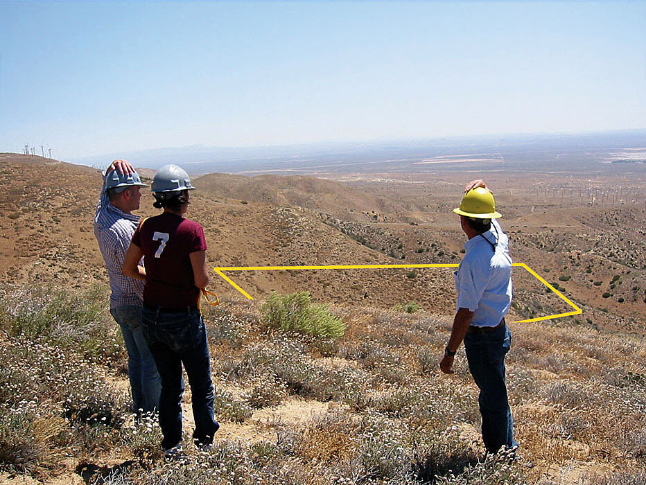 Three people wearing hard hats stand on a dry hillside, looking at a marked rectangular area below.
