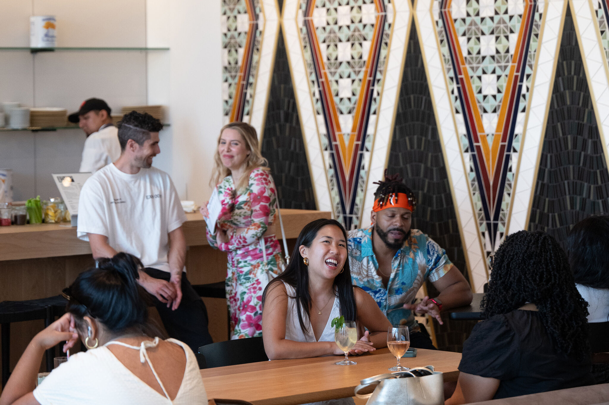 People enjoying lively conversation in a modern cafe with geometric art on the wall. Some are seated at tables, while others stand near the counter.