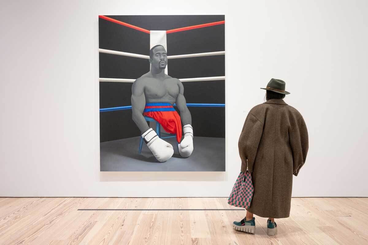 Person in large brown coat and hat stands in front of a large painting. The painting portrays a boxer without legs atop a stool in the center of a boxing ring.