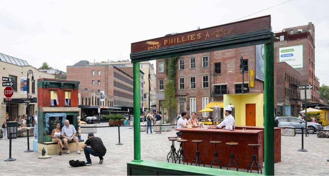 People interact with life-sized art installations, including a diner counter labeled "PHILLIES," in a busy city square.