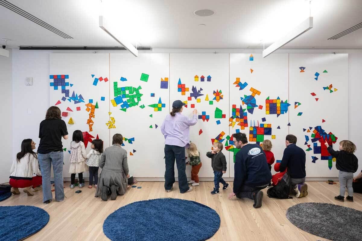A group of adults and children are gathered in a brightly lit room at the Whitney Museum, engaging in a creative activity with colorful magnetic geometric shapes. The shapes are arranged on a large white wall, forming a vibrant mosaic. Some participants are standing, while others are kneeling or sitting on the floor. The room has a clean, modern design with round blue and gray rugs scattered on the wooden floor. The atmosphere appears lively and collaborative.