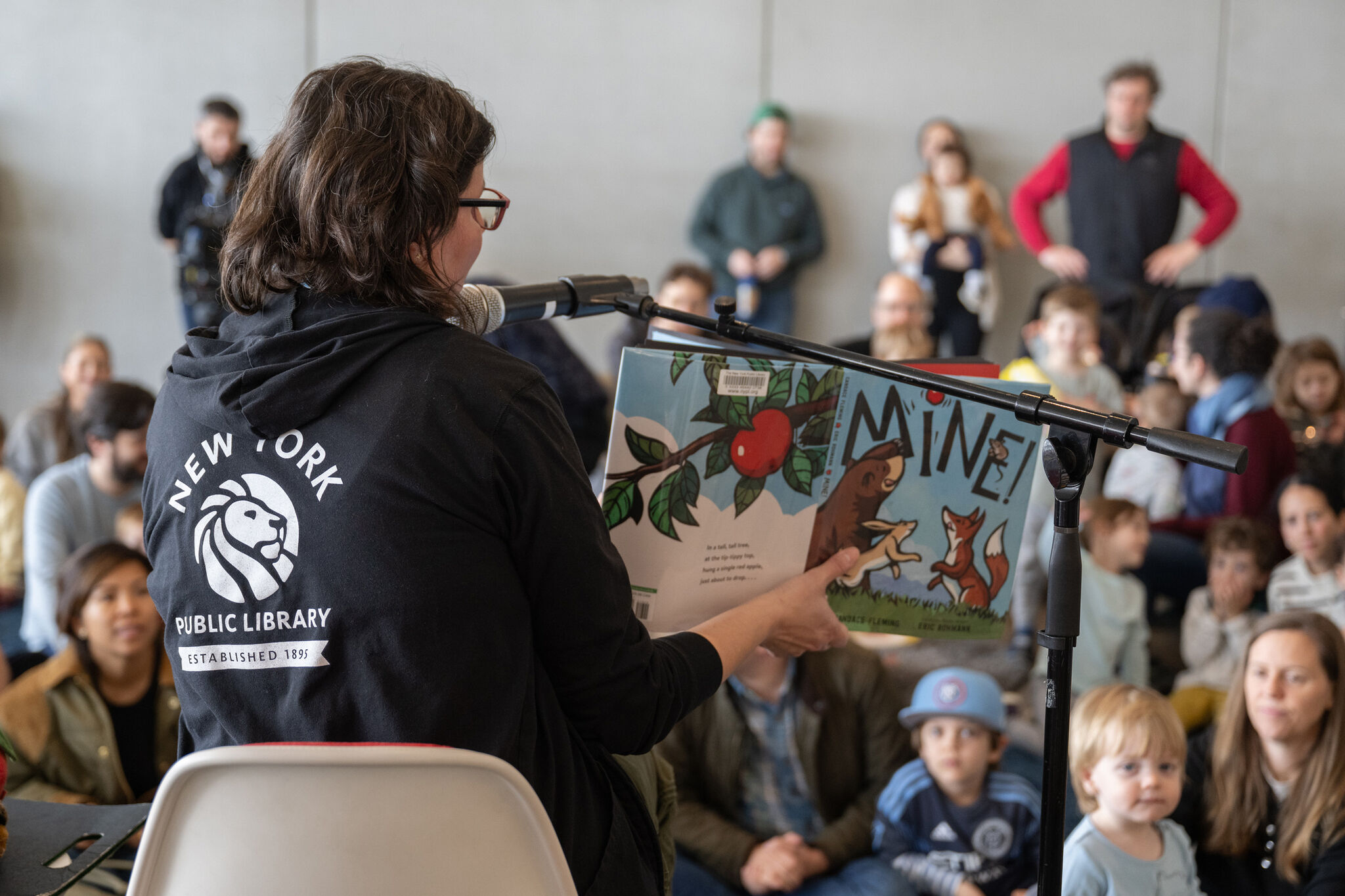A librarian reads the children’s book “Mine!” to a group of kids and parents at an event.