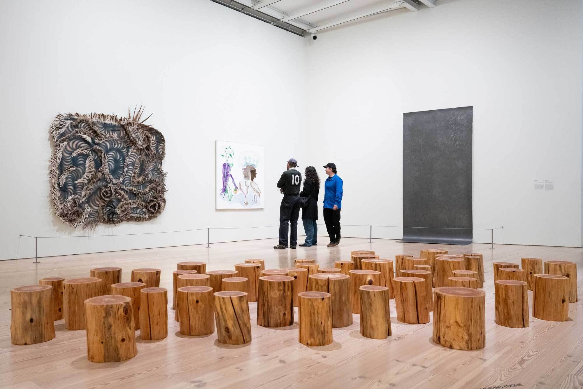 Three visitors observe art in a gallery. At the foreground is Maya Lin's art installation comprised of scattered wood logs; other artworks of mixed media are seen hung on adjacent walls.