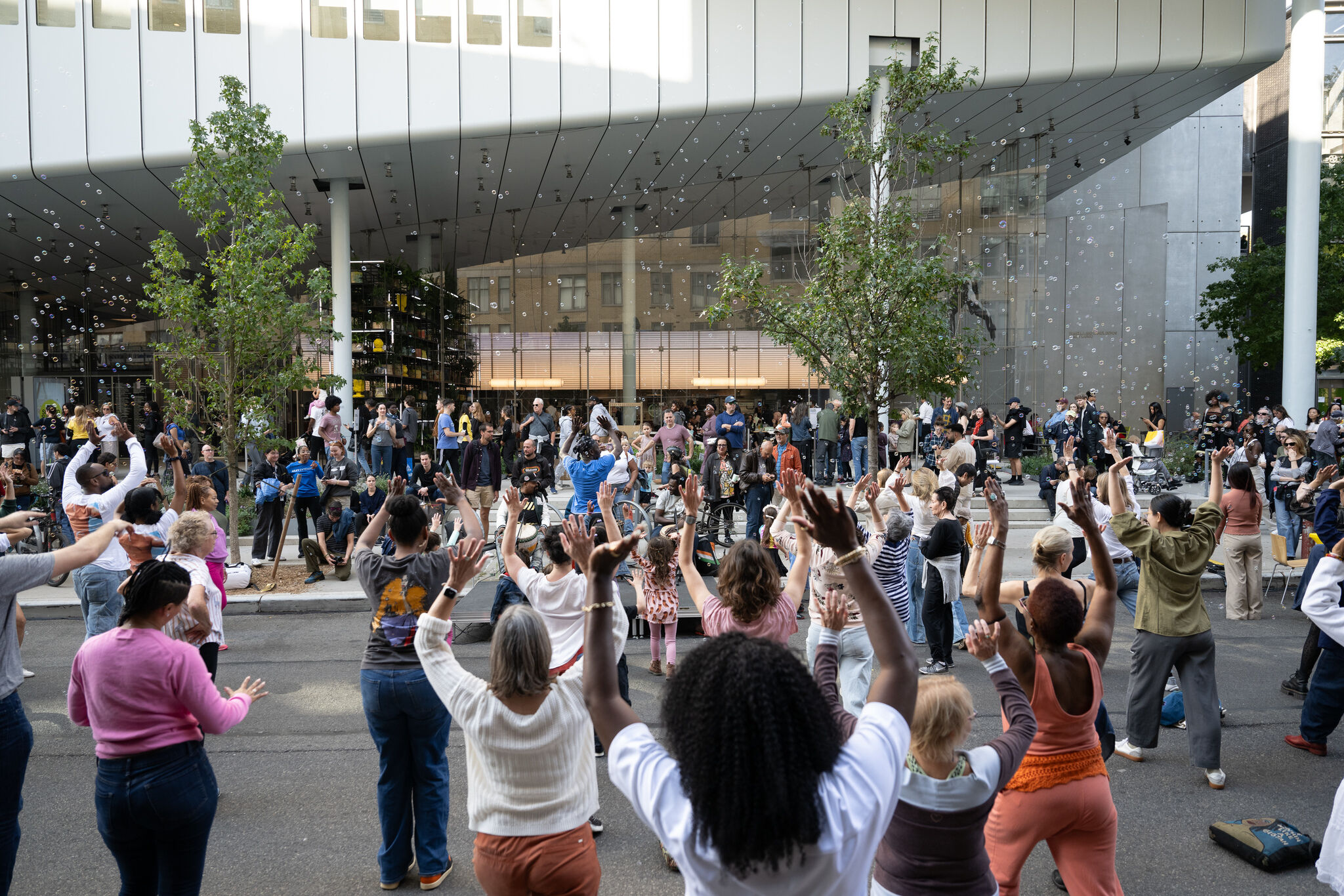 A large group of people joyfully dance with raised hands outside a modern building.