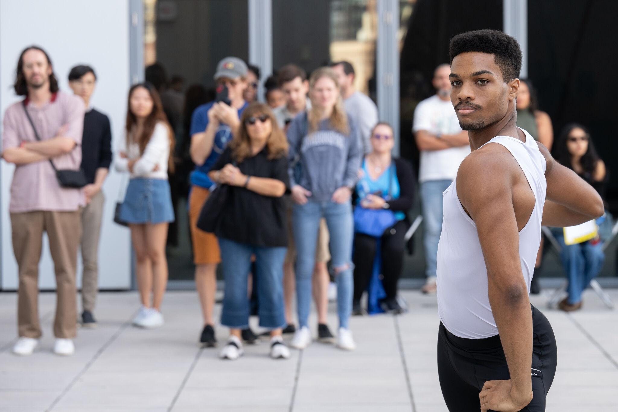 A dancer in a white tank top performs outdoors while a group of people watches in the background.