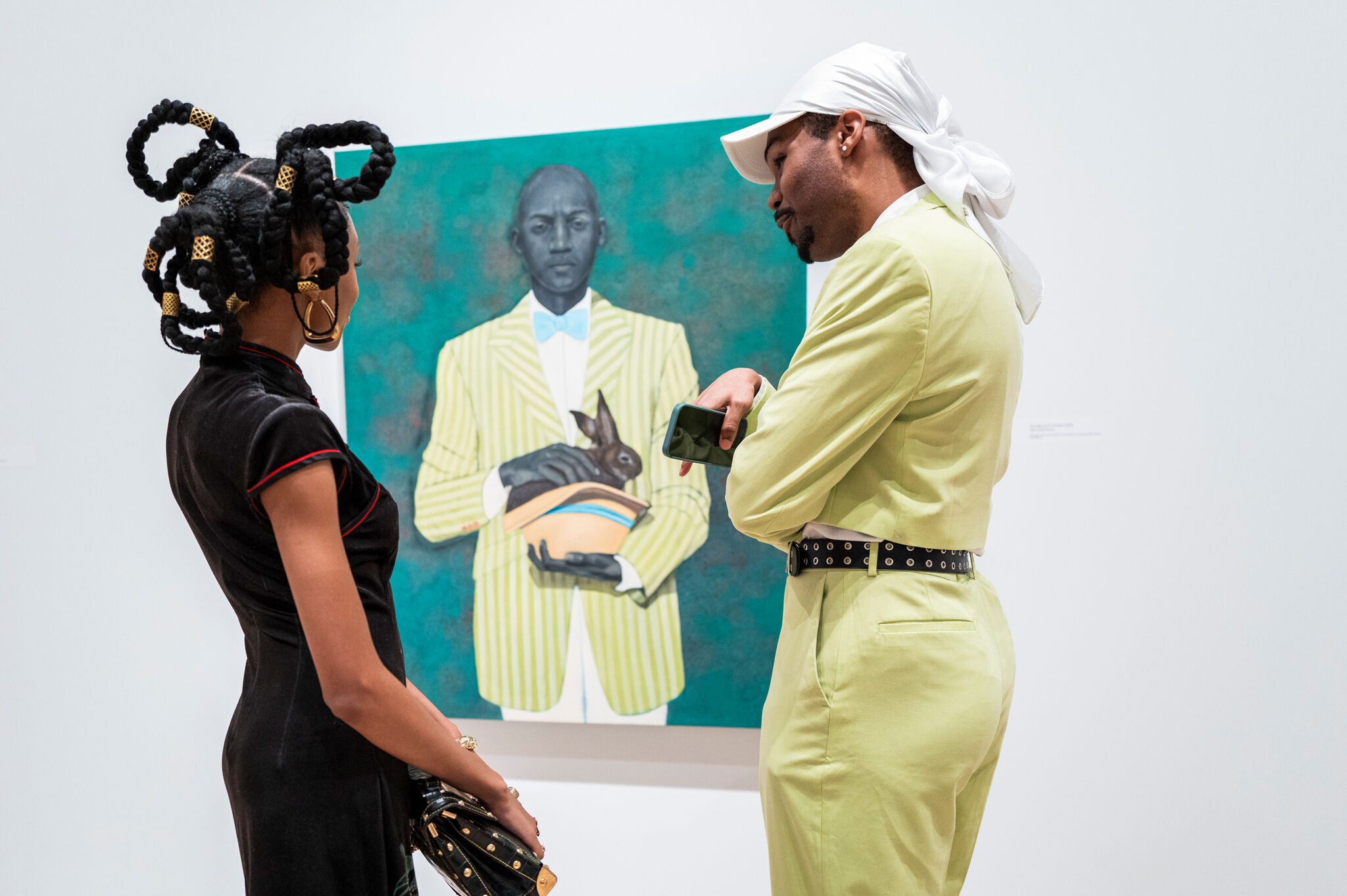 Two people stand in front of a painting of a man in a yellow suit holding a rabbit.