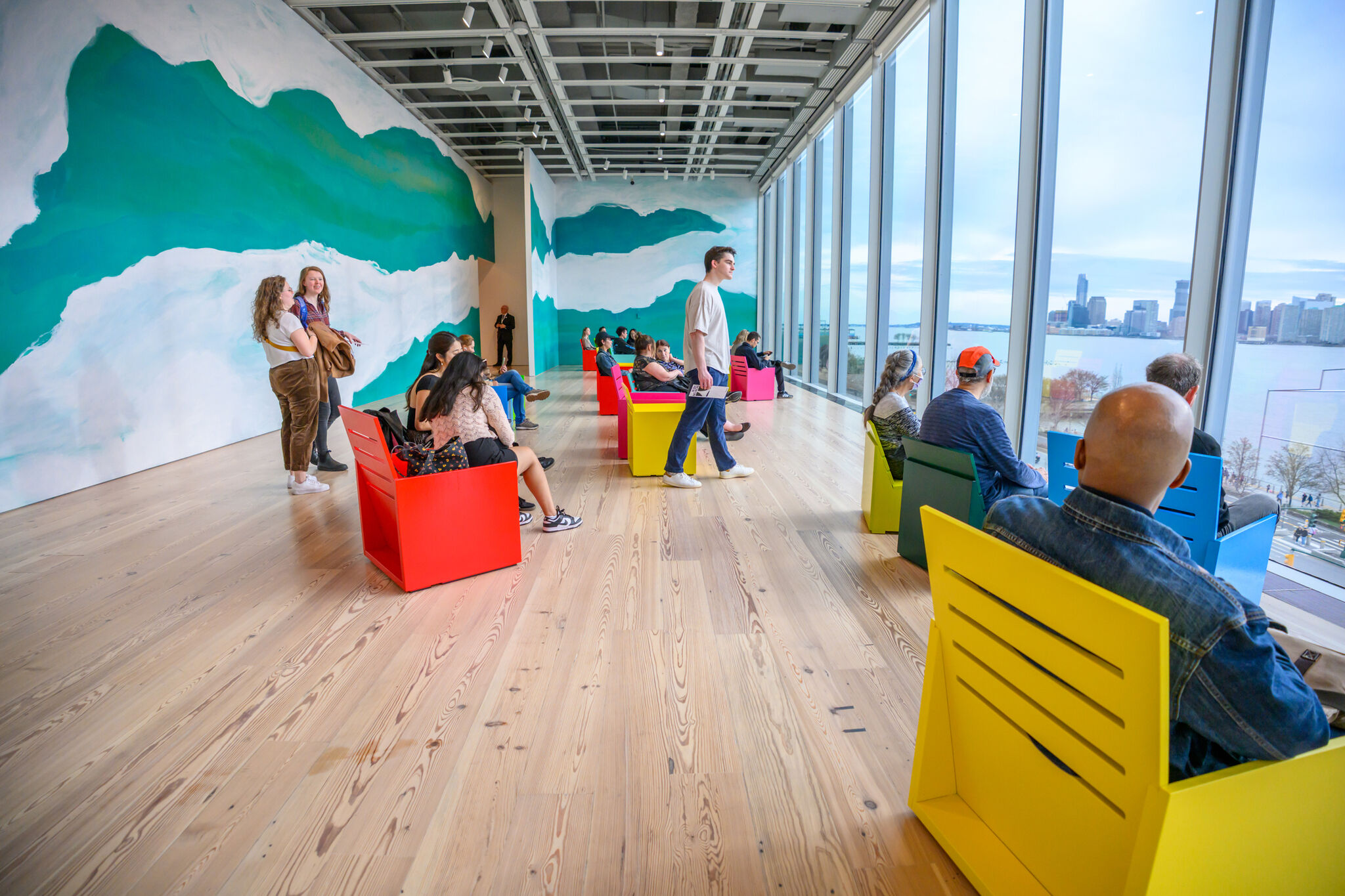 People sit in colorful chairs by large windows, looking out at a city skyline and river.