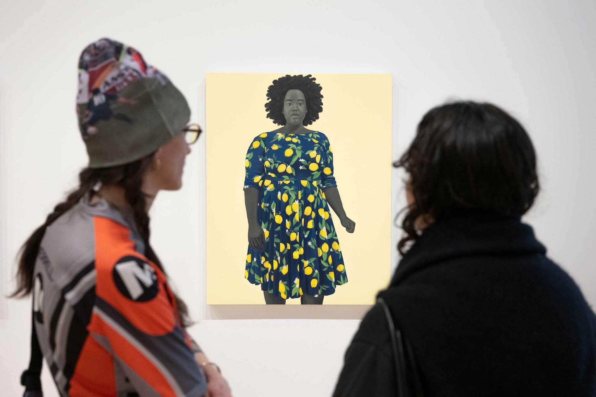 An image of two people in front of a portrait of a Black woman against yellow background. The person on the left is wearing a beanie and an orange shirt and the person on the right has long dark hair and a black shirt