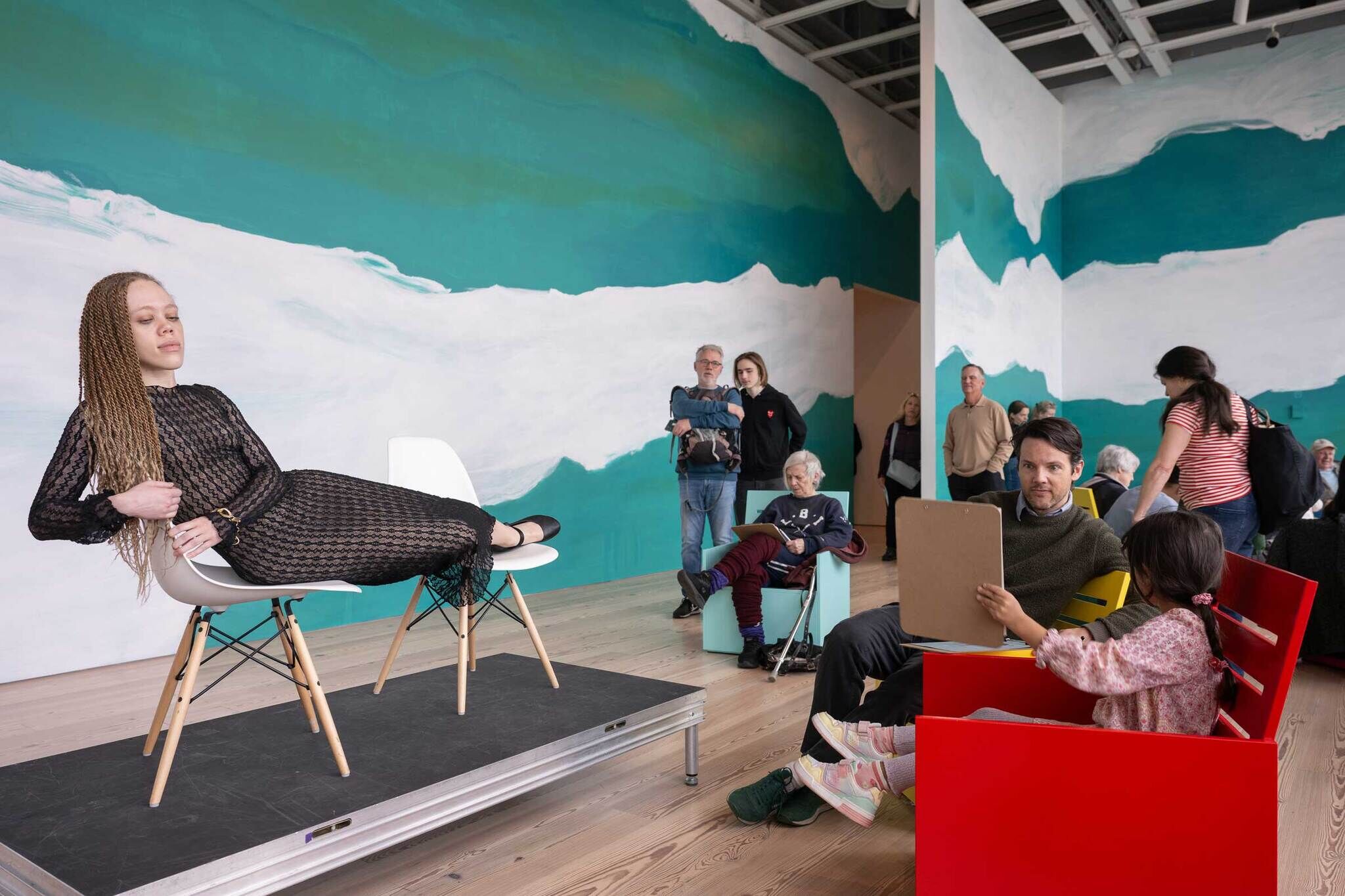 A woman in a black lace dress with long braids poses reclining on two white chairs on a small stage, serving as a live model for a figure drawing session. In the foreground, a young kid in pink holds a clipboard and draws, seated next to a grownup who admires the drawing. Around them, people of various ages observe or participate. The event is part of Free Second Sunday at The Whitney Museum of American Art.