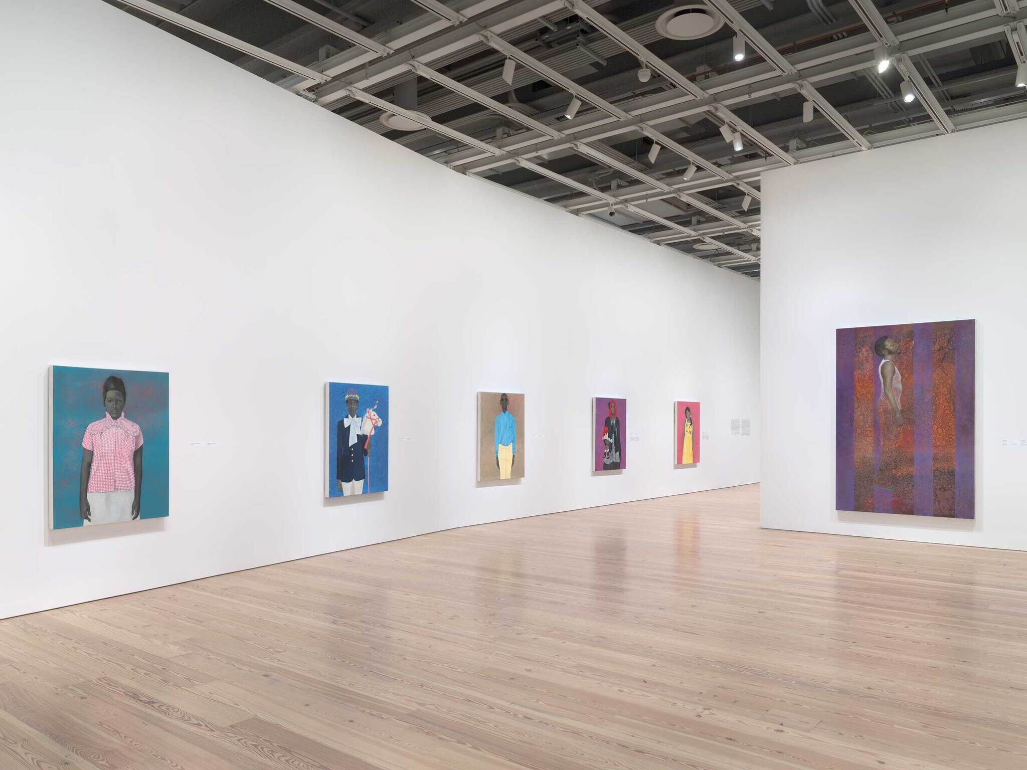 Several colorful paintings of people hang on white gallery walls with a wooden floor and open ceiling above.