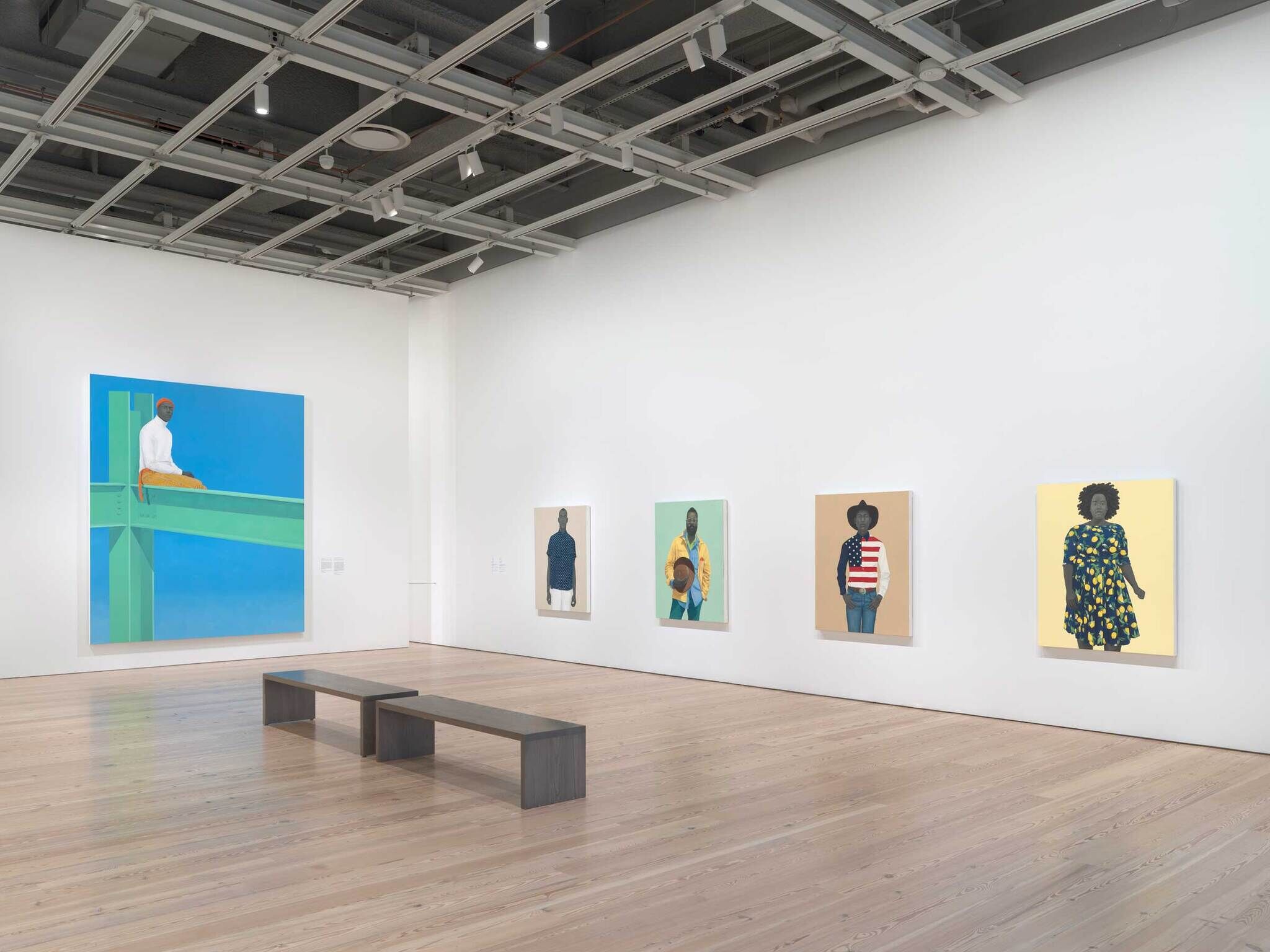 Art gallery with five colorful portraits of people on white walls and two simple benches on a wooden floor.