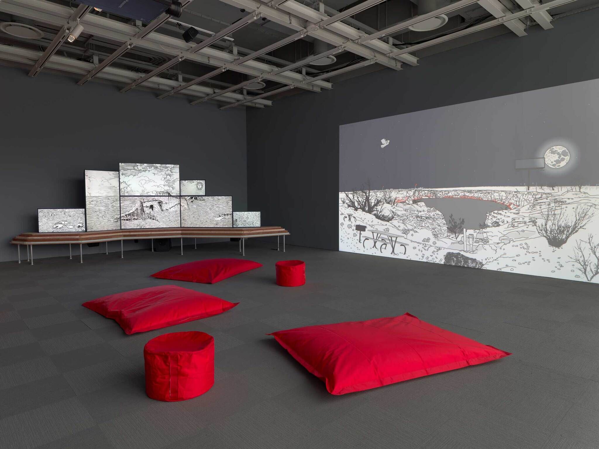 Several red cushions and stools face a wall with a large animated landscape projection and multiple screens showing similar art.