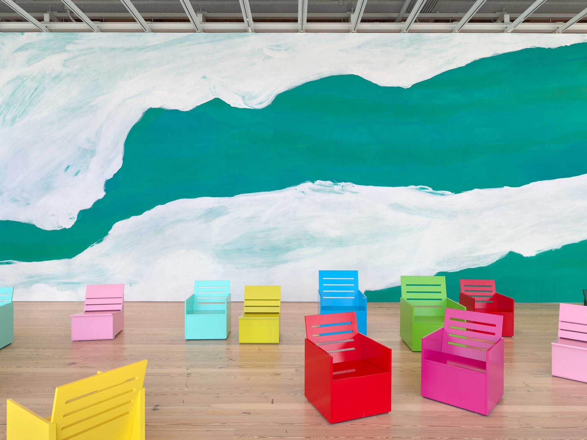 Brightly colored chairs are arranged on a wooden floor in front of a large abstract green and white mural.