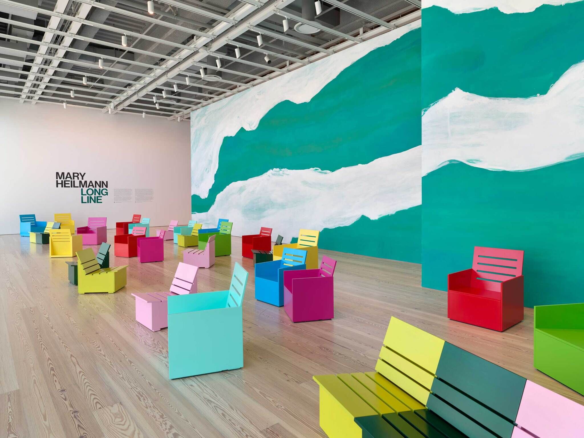 Room with colorful box-shaped chairs, a large green and white mural, and the text “Mary Heilmann Long Line” on the wall.