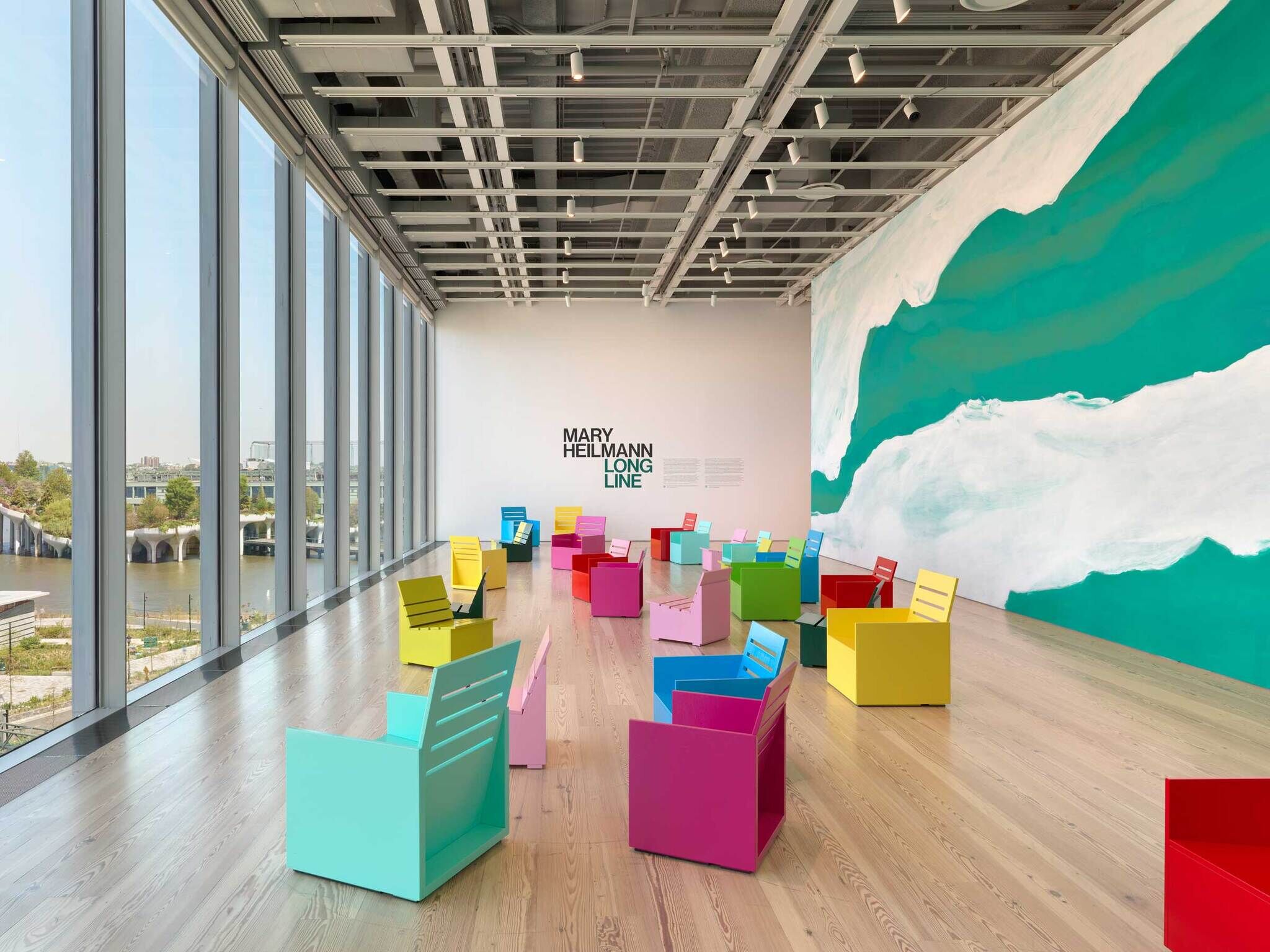 Brightly colored chairs are arranged in a spacious gallery with large windows and a wall reading “Mary Heilmann Long Line.”