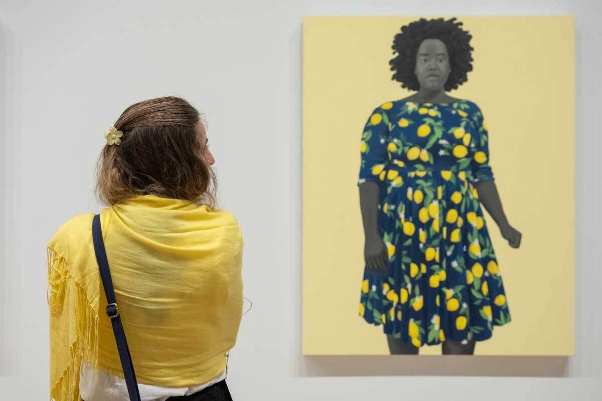 A woman with blonde hair in a yellow shawl is looking at a painting of a woman wearing a lemon-print dress.