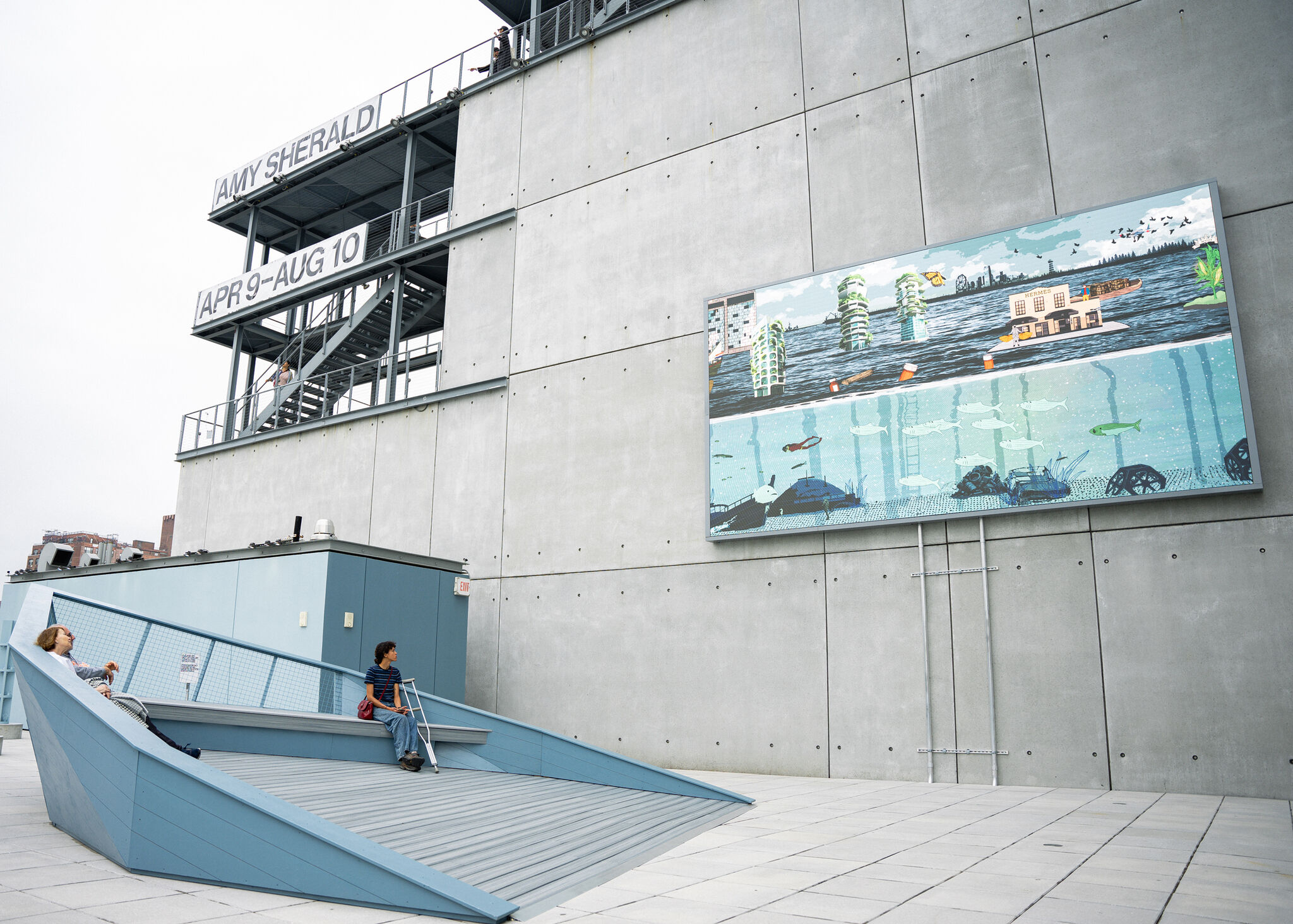 Two people sit on a blue bench outside, looking at a large mural on a concrete building wall. Signs read “AMY SHERALD” and “APR 9–AUG 10.”
