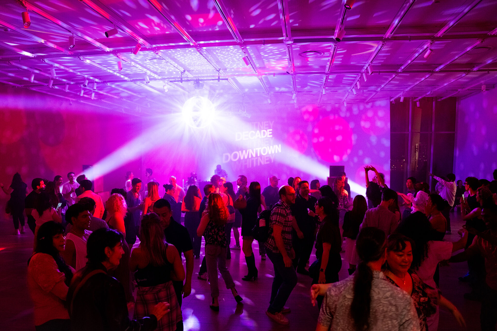 A large group of people dance under purple lights, with “DECADE DOWNTOWN WHITNEY” projected on the wall.
