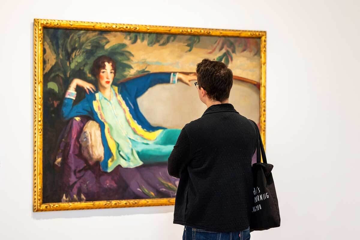 A person stands in front of a large, colorful portrait painting in an art gallery. The painting depicts Gertrude Vanderbilt Whitney in vibrant clothing, including a turquoise dress and a blue robe with yellow accents, lounging on a purple couch with one arm draped over the backrest.