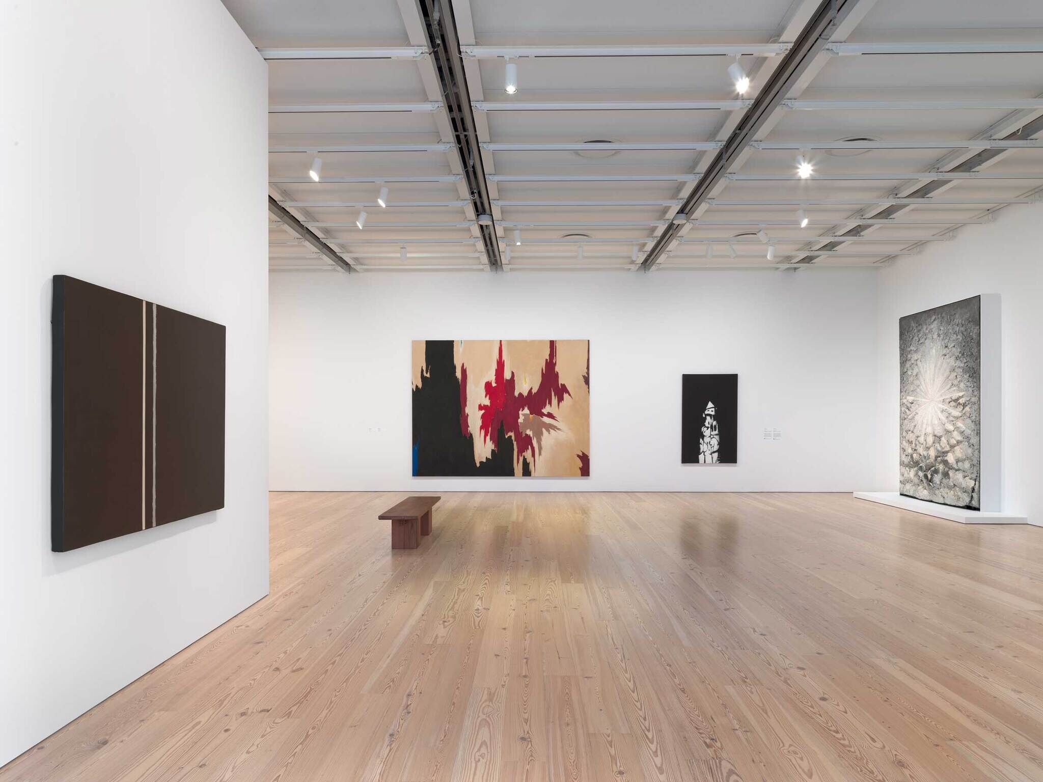 Art gallery with abstract paintings on white walls, wooden floor, and a wooden bench in the center.