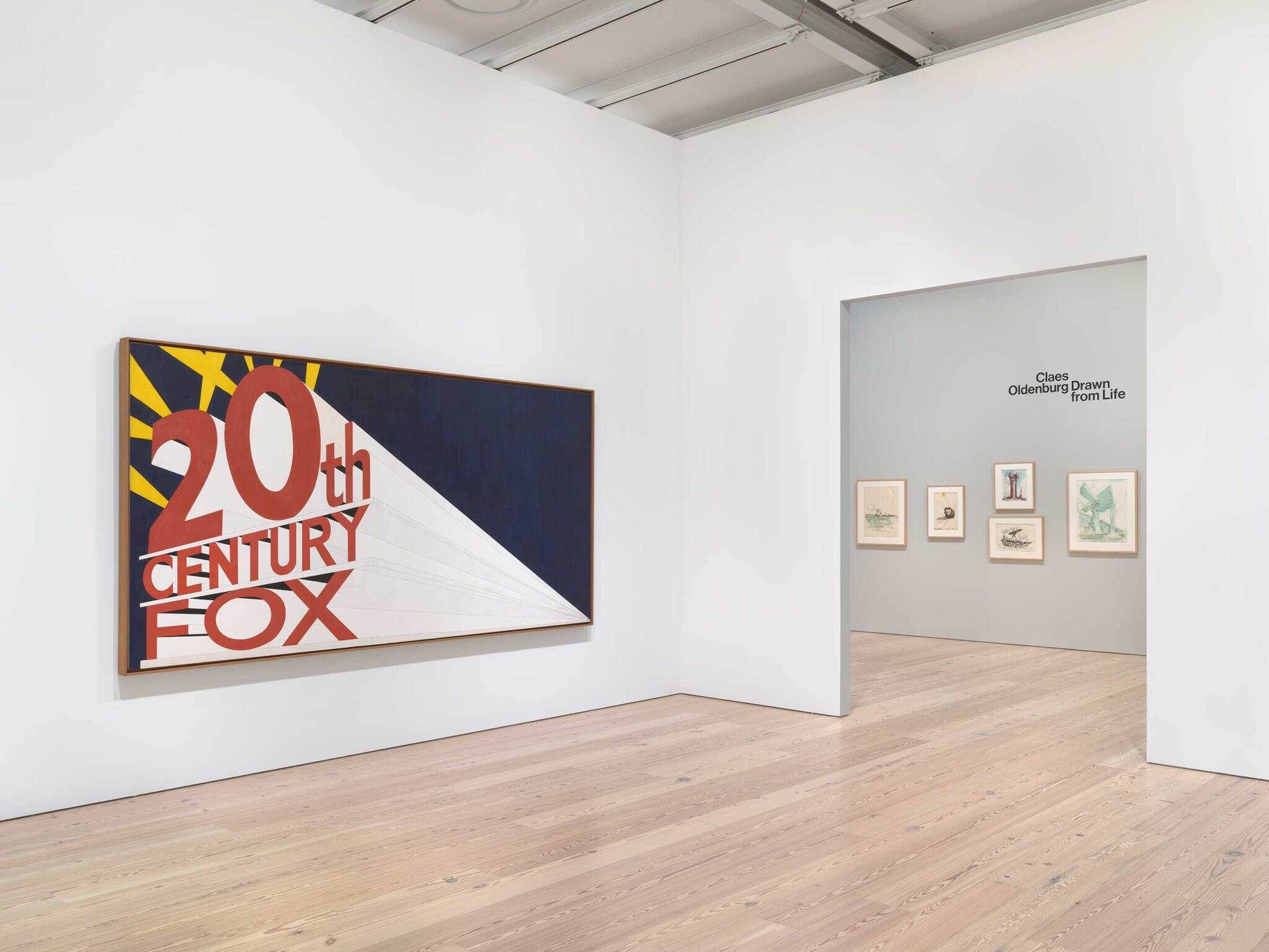 An art gallery with a big pop art painting on the left and on the right a window into a smaller gallery with colorful sketches framed.