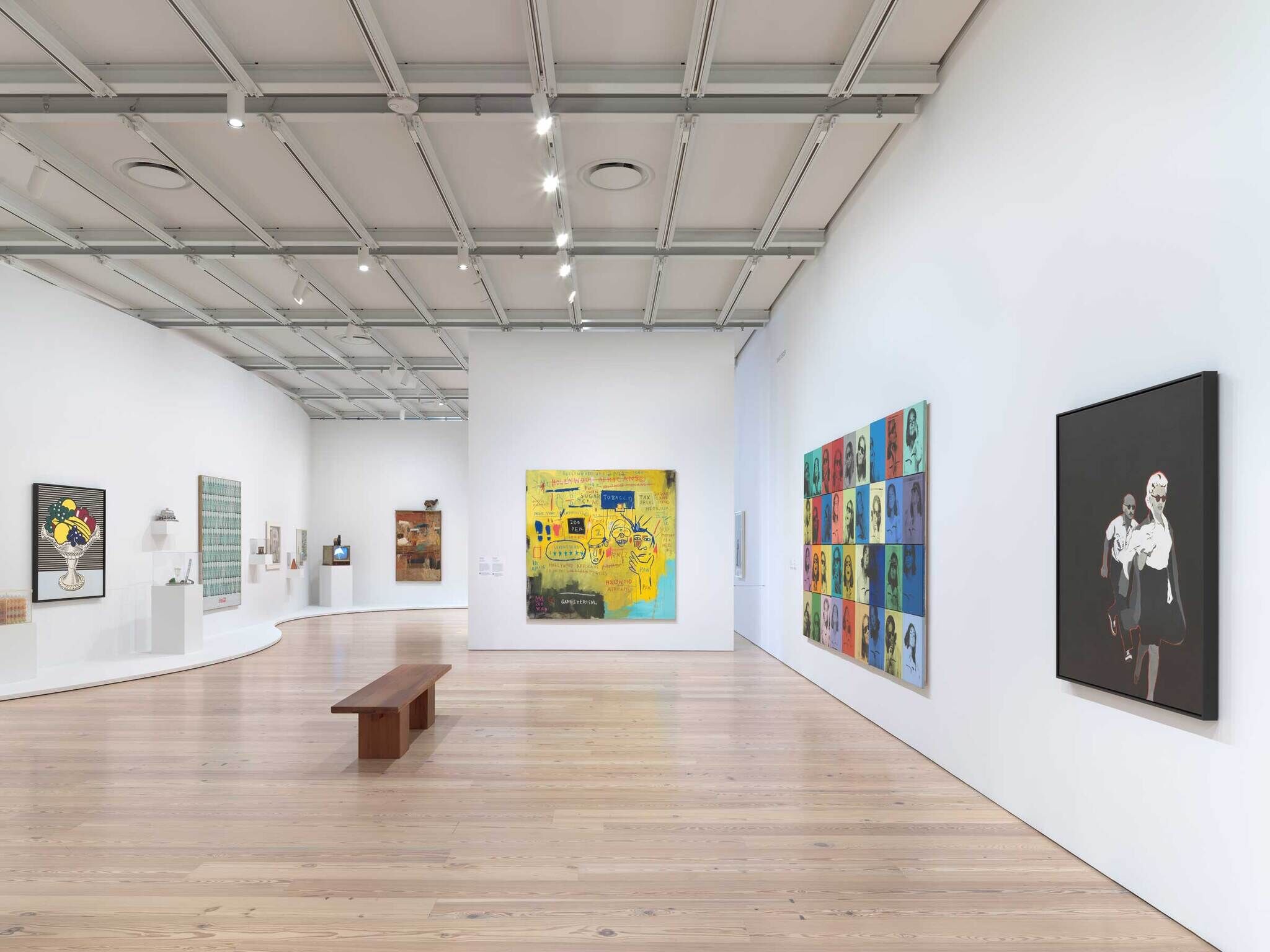 Modern art gallery with colorful paintings on white walls, wooden bench in the center, and bright overhead lighting.