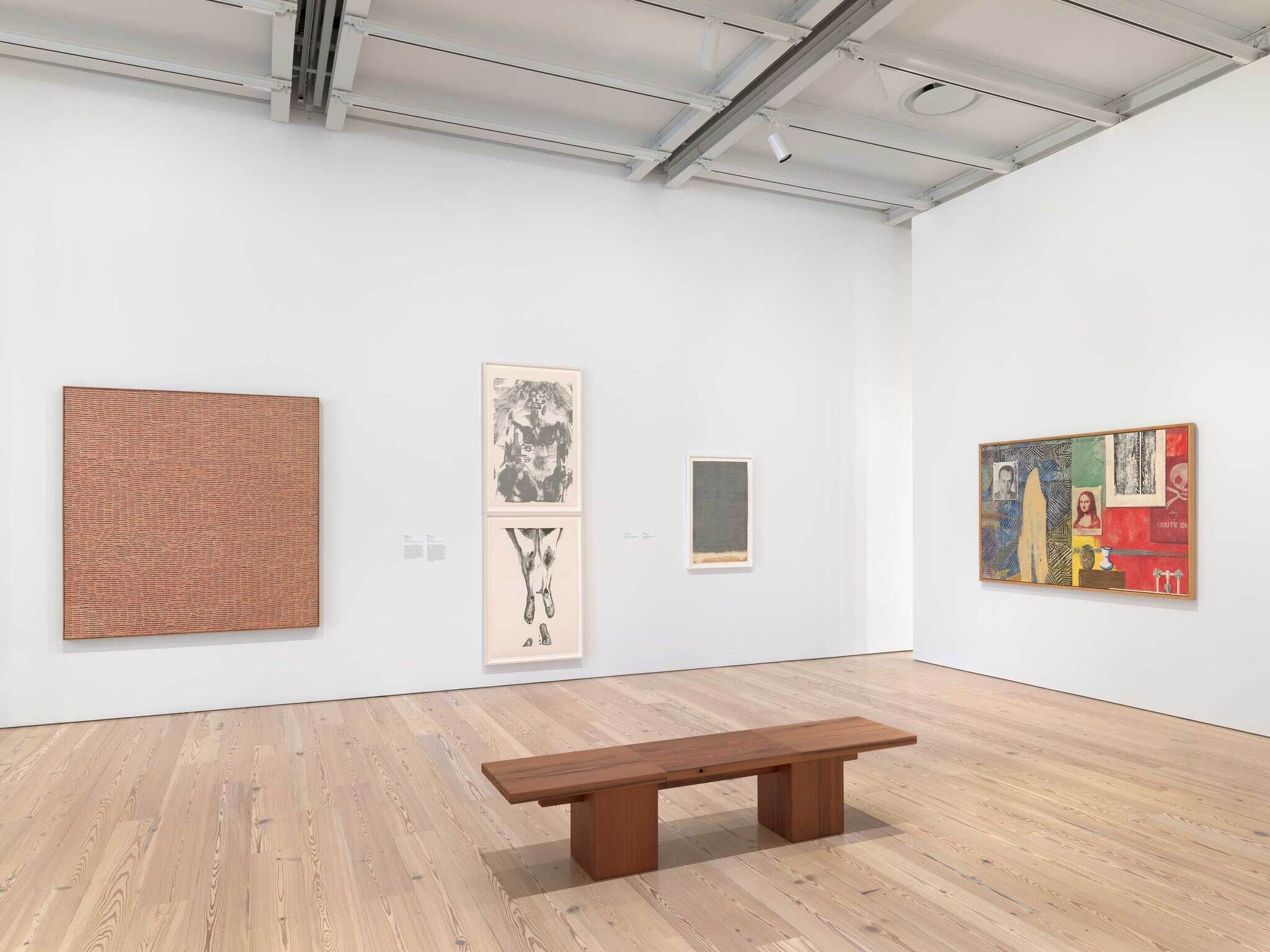 Art gallery room with several modern paintings on white walls and a wooden bench in the center.