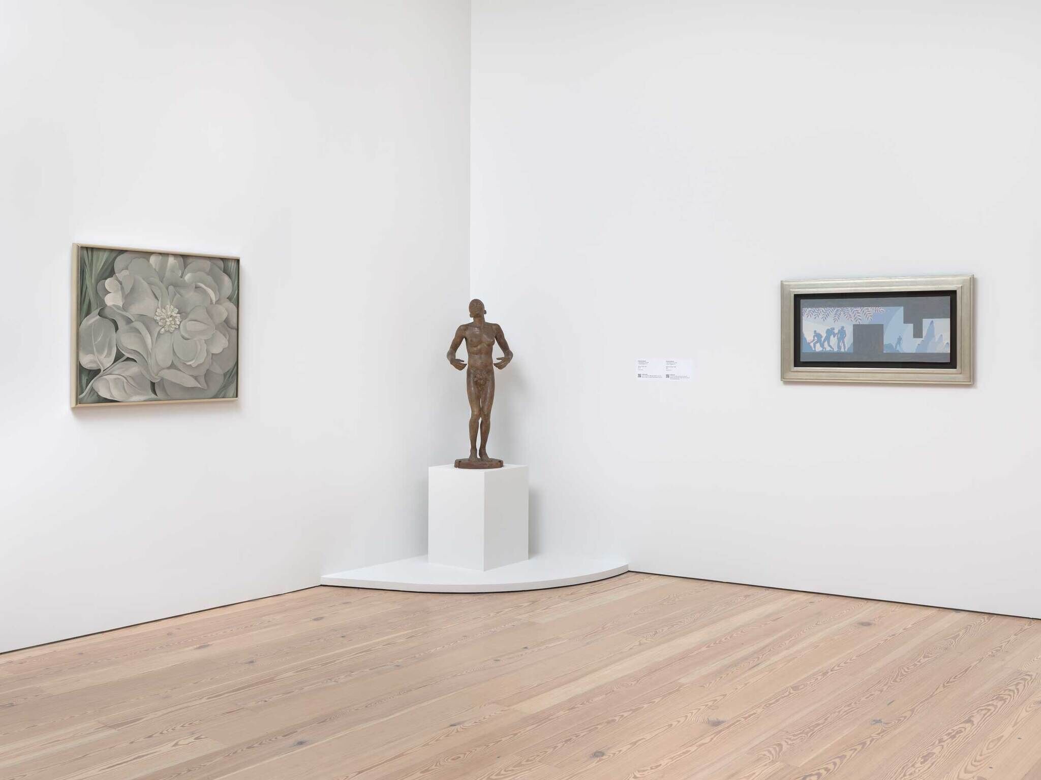 From left to right in an art gallery with white walls and a light tan floor, a painting of a white flower, a brown human sculpture, and a blue framed horizontal painting