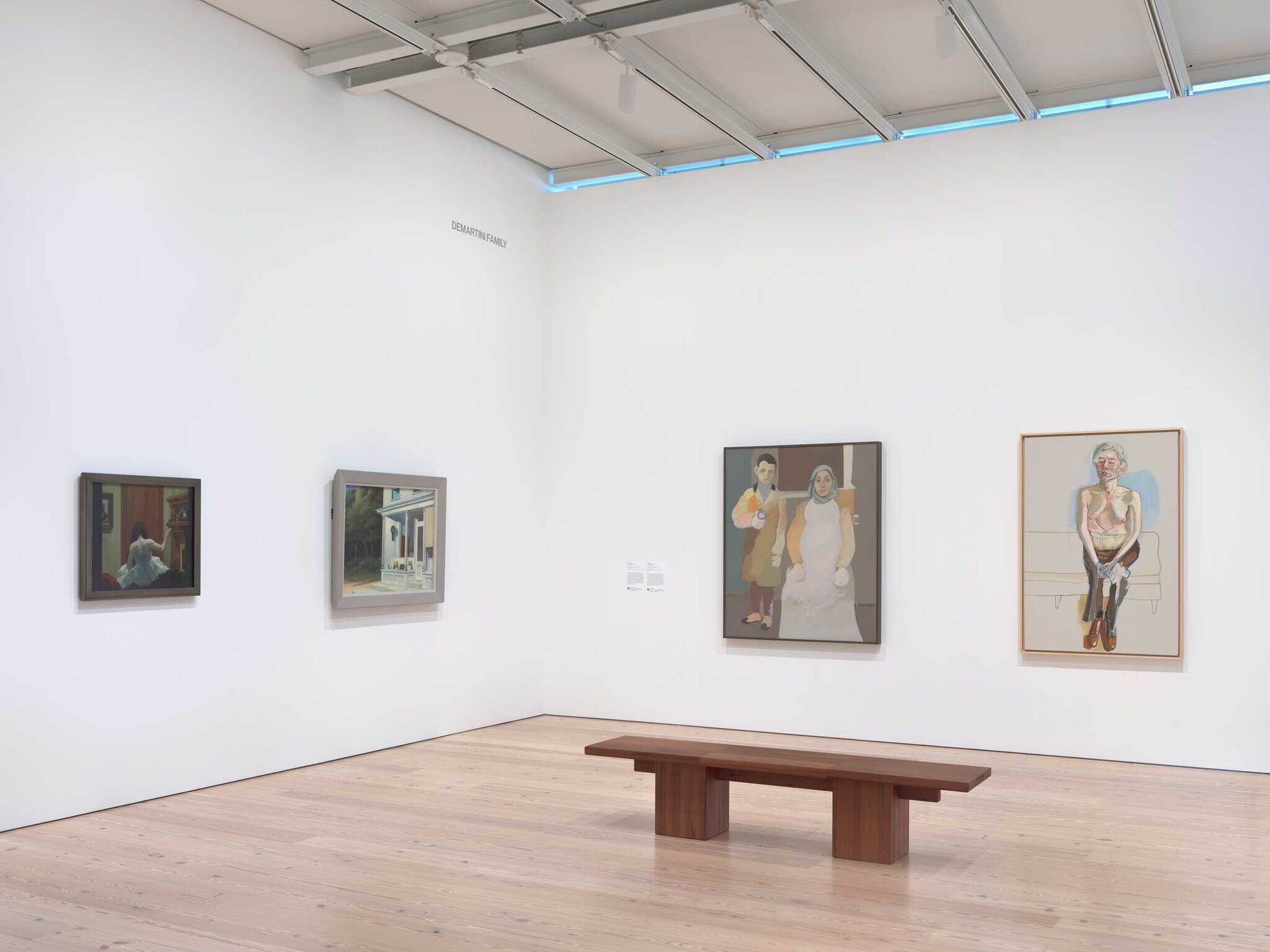 Four paintings hang on a white gallery wall above a wooden bench.