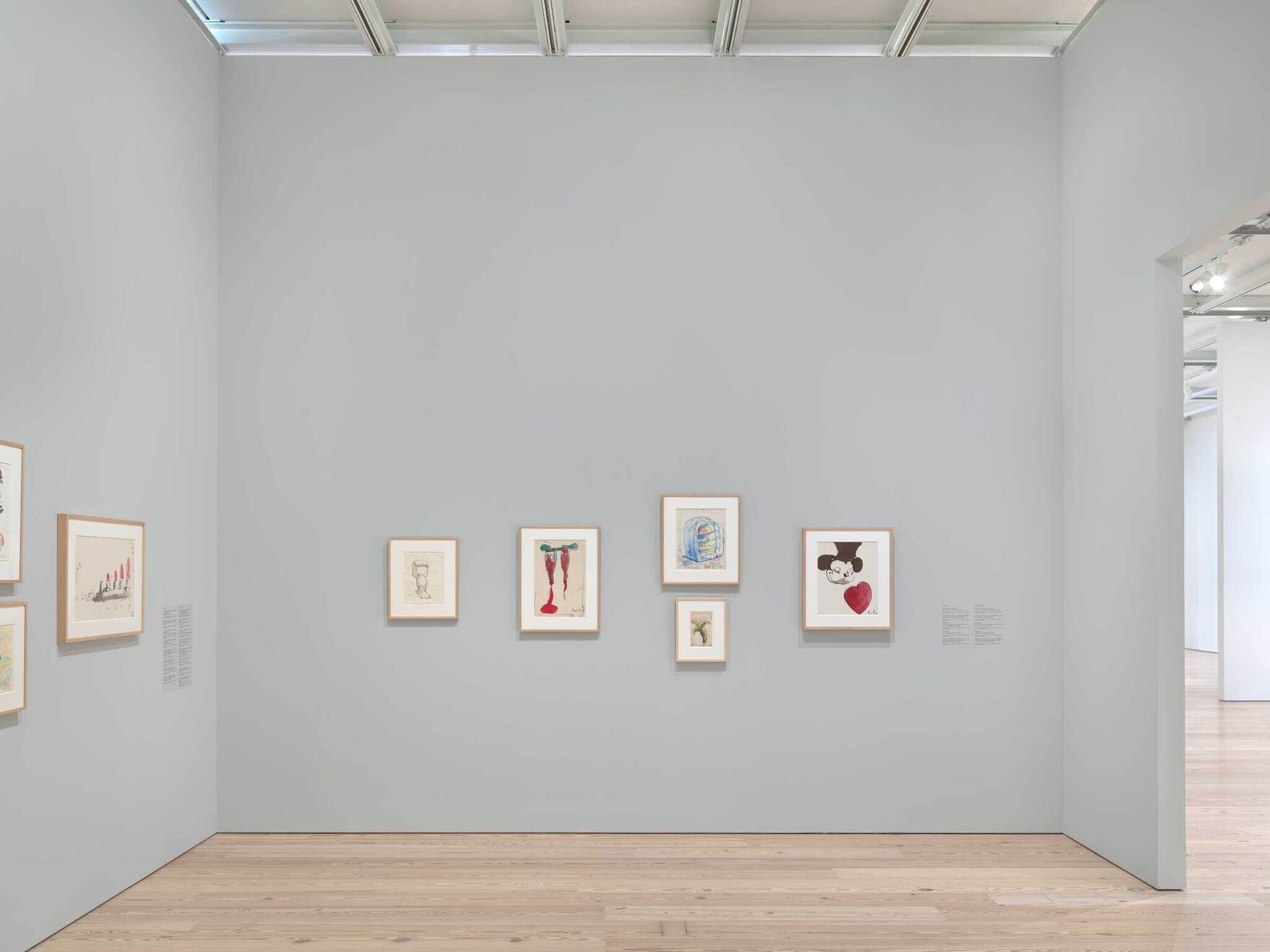 Five framed drawings hang on a light gray gallery wall, each showing different abstract or cartoon-like figures.